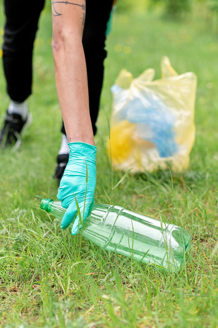 A gloved hand reaching down into the grass to pick up a discarded clear plastic bottle, a bag of collected litter behind it.