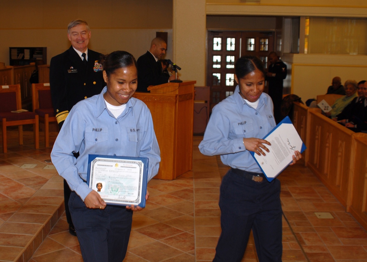 Two identical twin women in matching U.S. Navy uniforms, side by side, holding certificates.