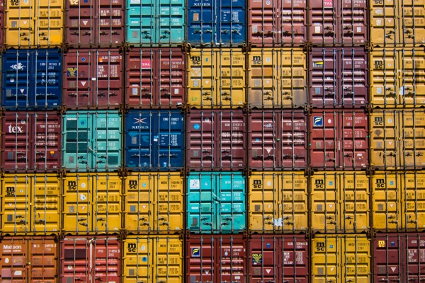 An overhead grid of brightly colored stacked shipping container ends, yellow, rust red, and teal, packed in orderly rows.
